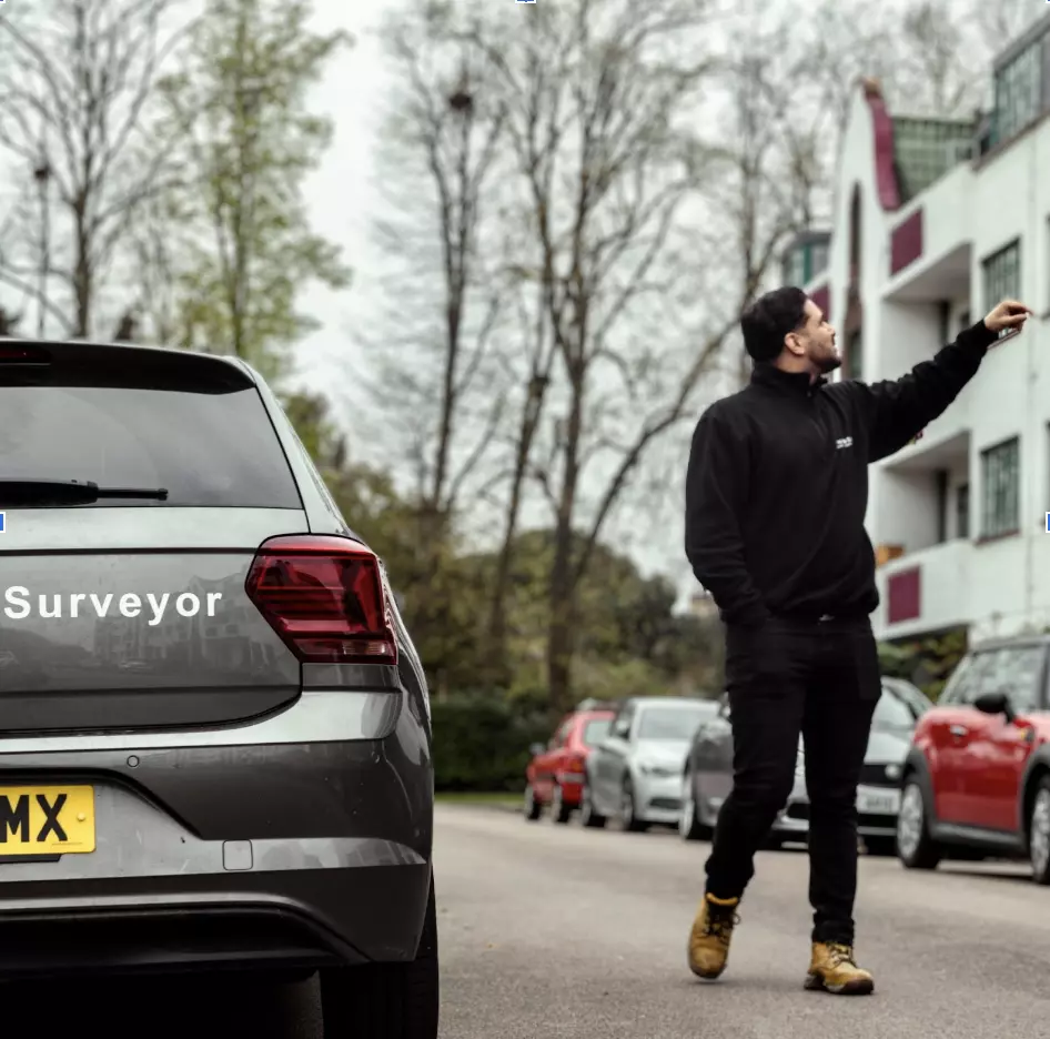 Surveyor pointing towards a building, parked car with "Surveyor" branding, residential street with various vehicles, context of property maintenance and repairs.