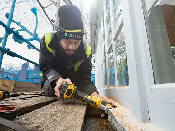 Worker using a power drill for sash window restoration on a building, showcasing expert craftsmanship and attention to detail in preserving period home aesthetics.