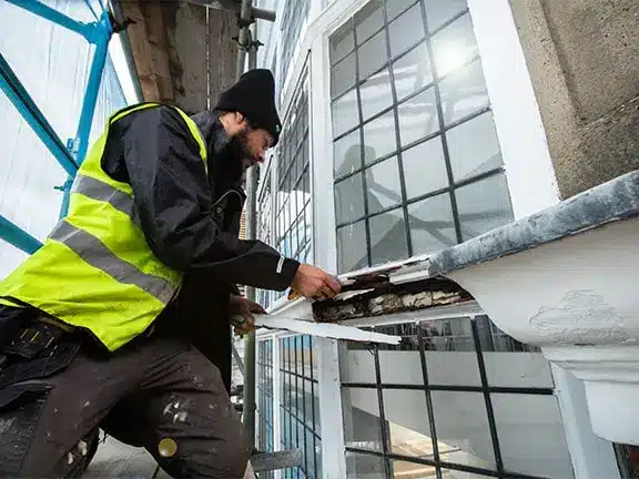Worker in safety gear repairing a sash window, showcasing restoration techniques for period homes, with scaffolding in the background.