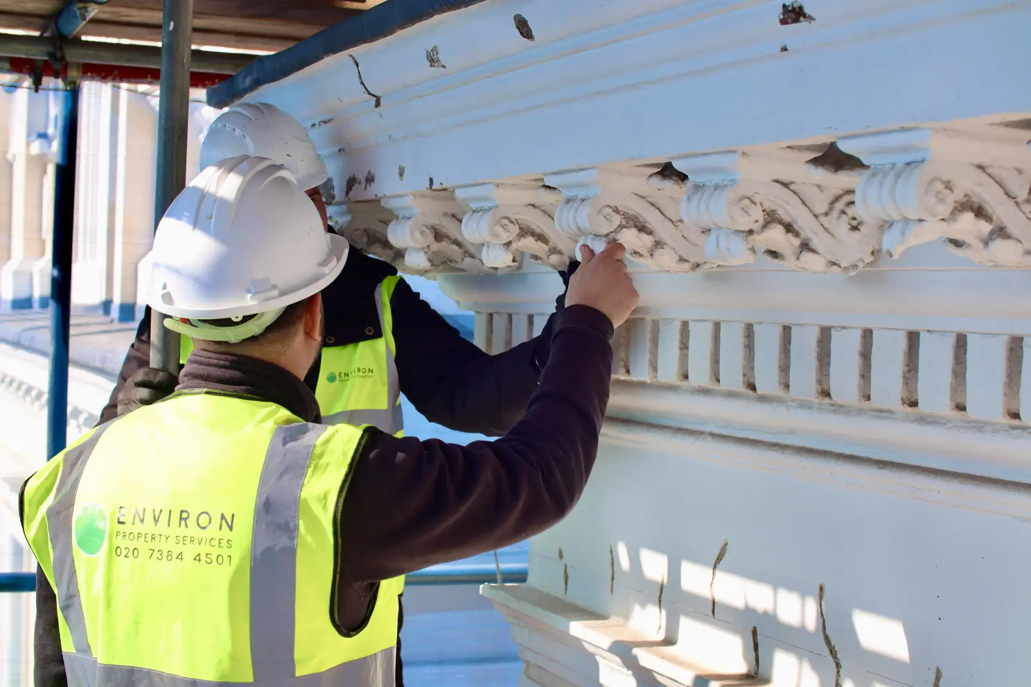 Two workers in safety helmets and high-visibility vests from Environ Property Services, performing stone masonry cleaning and restoration on a decorative architectural feature.