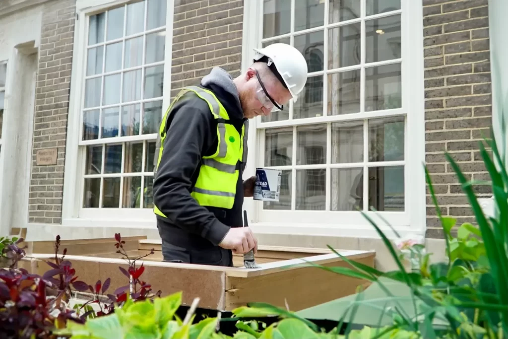 Worker in safety gear repairing timber window frame outside period property, showcasing expertise in restoration and preservation of historical architecture.
