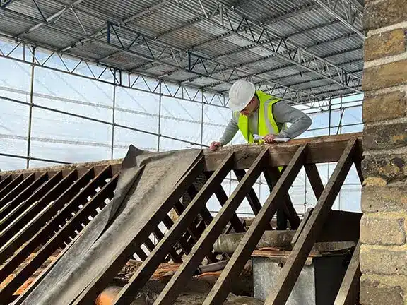Worker in safety gear inspecting wooden beams during stone wall restoration process, showcasing expertise in structural repairs.