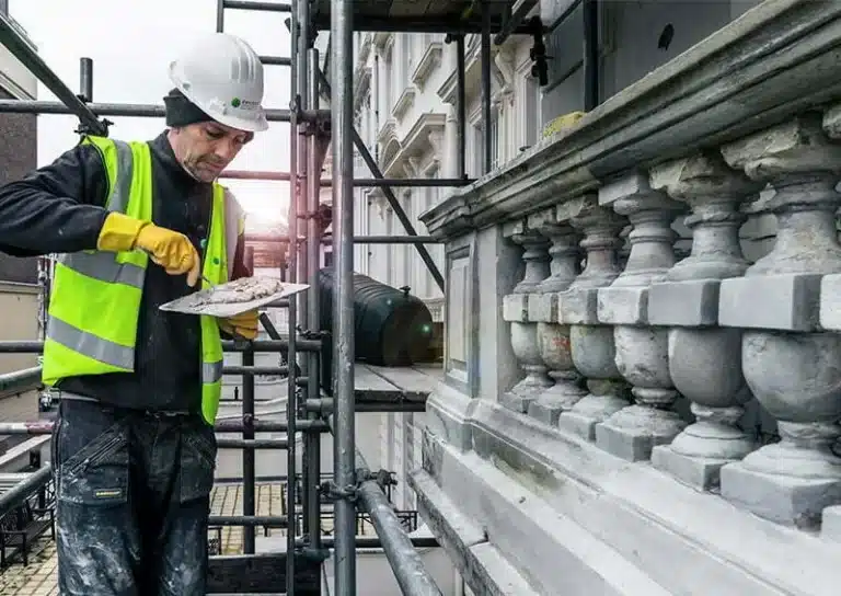 Worker in safety gear applying plaster to ornate stonework on scaffolding, showcasing restoration techniques for building maintenance.