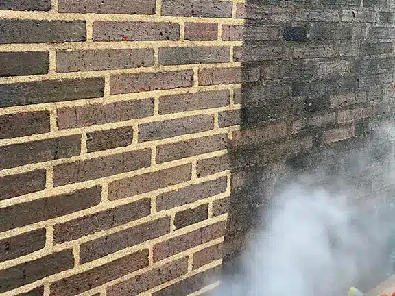 Brick cleaning process showcasing a partially restored brick wall, highlighting the contrast between cleaned and uncleaned sections, with steam rising from cleaning equipment.
