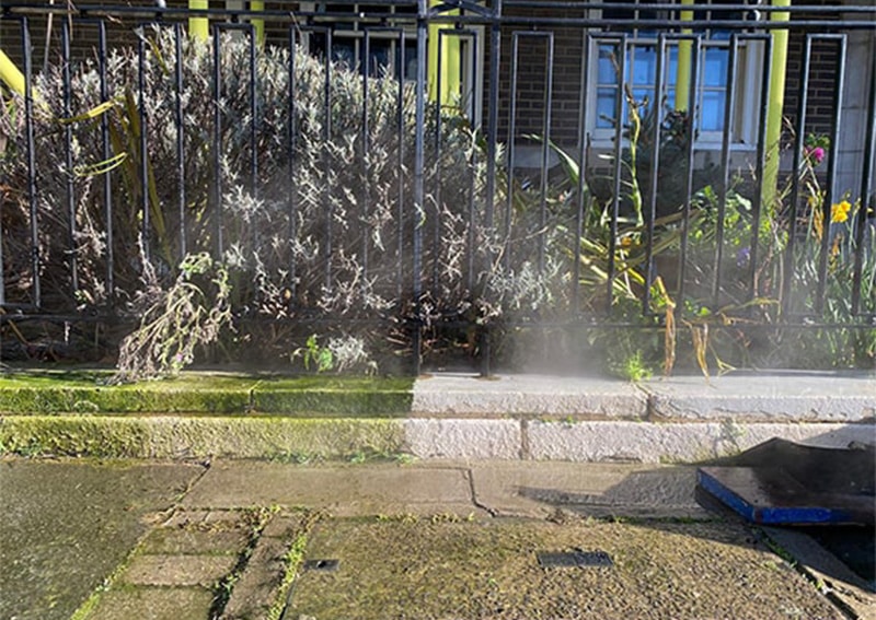 Driveway cleaning process showing high-pressure washing removing dirt and grime from stone paving, with surrounding greenery and fence in London.