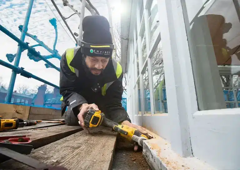 Skilled tradesman repairing sash window frame with power tools, focused on restoration work, scaffolding in background, showcasing expert craftsmanship in sash window repair services.