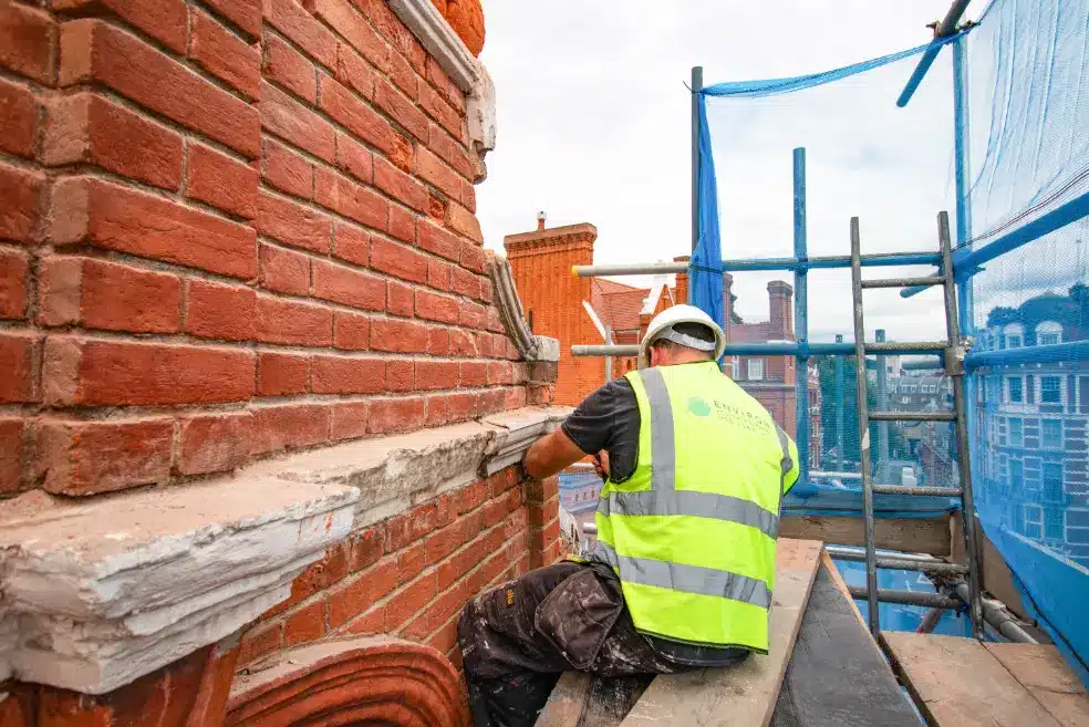 Brick repointing in progress, worker applying mortar to brick wall, wearing safety gear and high-visibility vest, scaffolding and blue protective netting in background, showcasing restoration services in London.