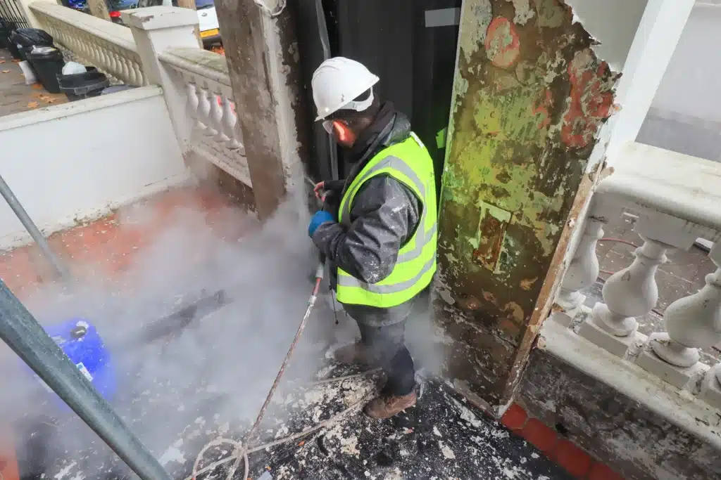 Worker in safety gear using a pressure washer to clean brickwork, steam and debris visible, showcasing professional brick cleaning services in London.