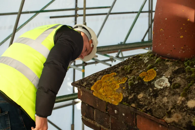 Surveyor inspecting a chimney with visible moss and lichen, wearing a hard hat and high-visibility vest, amidst scaffolding, highlighting restoration assessment for historic buildings.