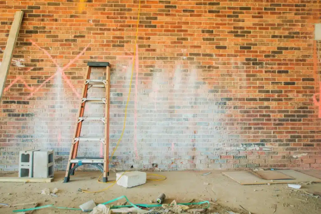 Brick wall with graffiti marks, ladder for access, construction debris, and cleaning supplies, illustrating preparation for effective graffiti removal techniques by Environ Restoration Services.