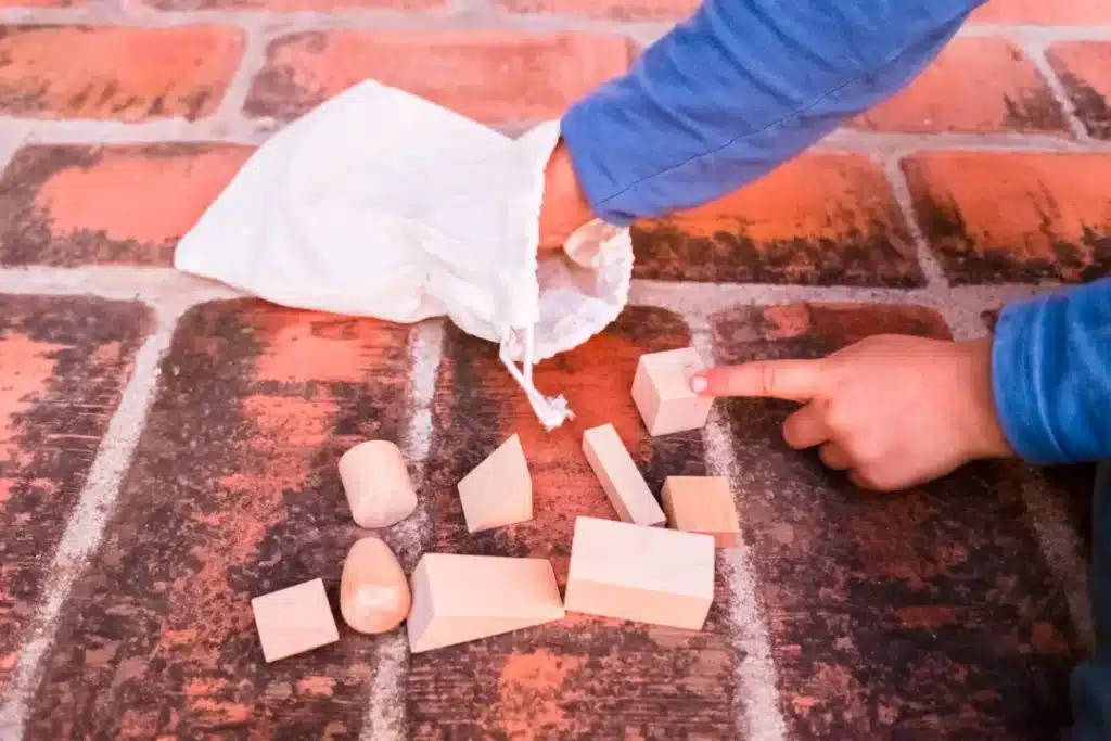 Child pointing at wooden building blocks on a brick surface, with a white bag nearby containing additional blocks, illustrating playful learning and creativity in a home environment.
