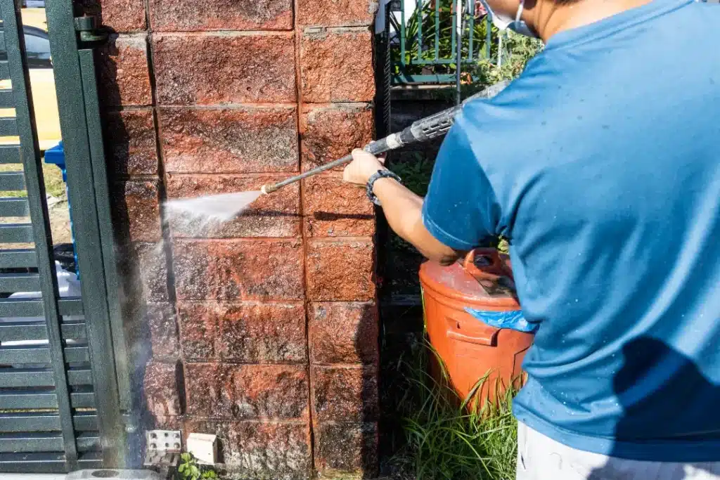 Person using a pressure washer to clean a brick wall, demonstrating safe practices for maintaining brick surfaces.