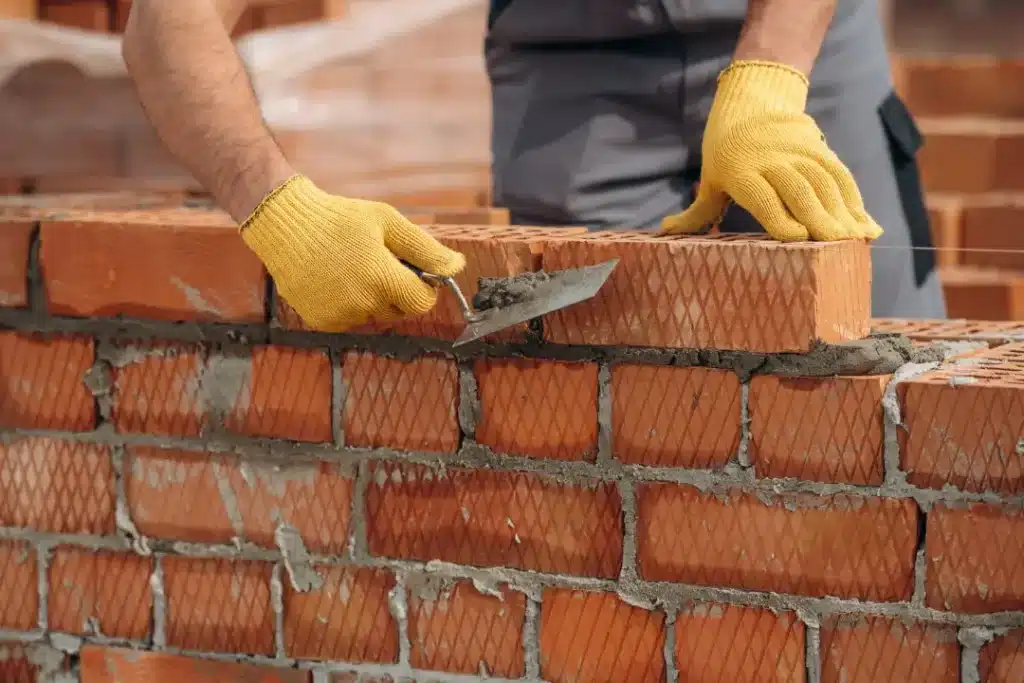 Person wearing yellow gloves laying bricks with trowel, demonstrating brickwork techniques relevant to cleaning and maintenance.