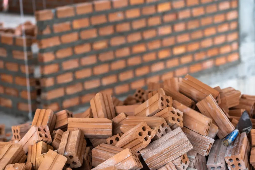 Piled bricks showcasing various shapes and sizes, with a brick wall under construction in the background, illustrating key characteristics of good-quality bricks for construction projects.