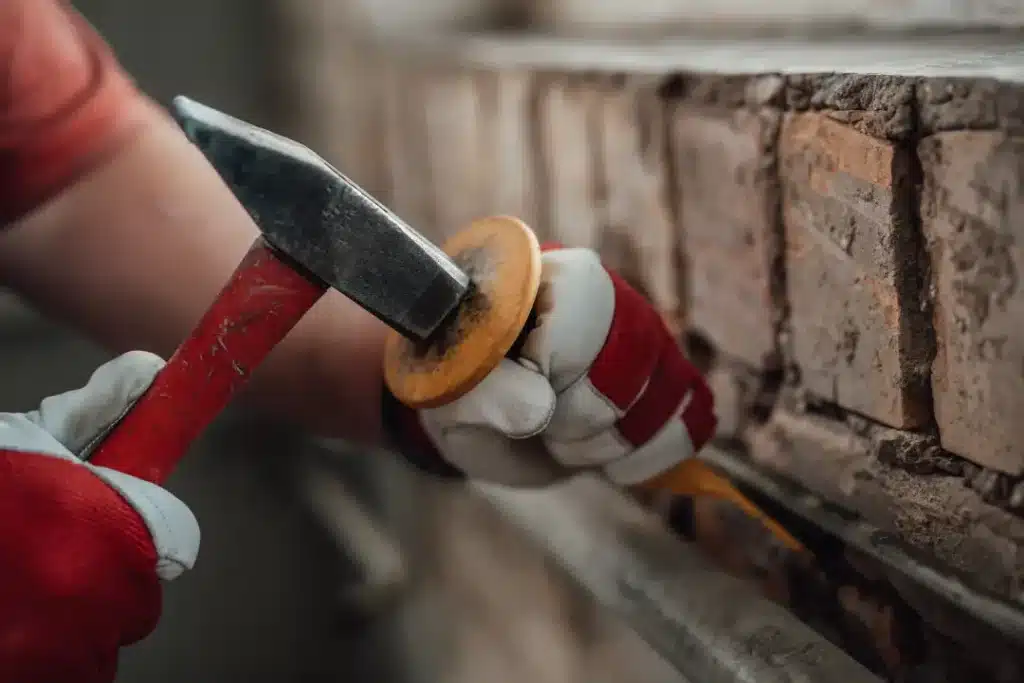 Close-up of a person wearing gloves using a hammer and chisel to repoint brickwork, illustrating professional brick restoration techniques relevant to maintaining historic buildings.