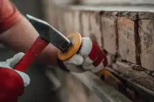 Person using a hammer and chisel to remove old mortar from brickwork, demonstrating the repointing process relevant to brick restoration.