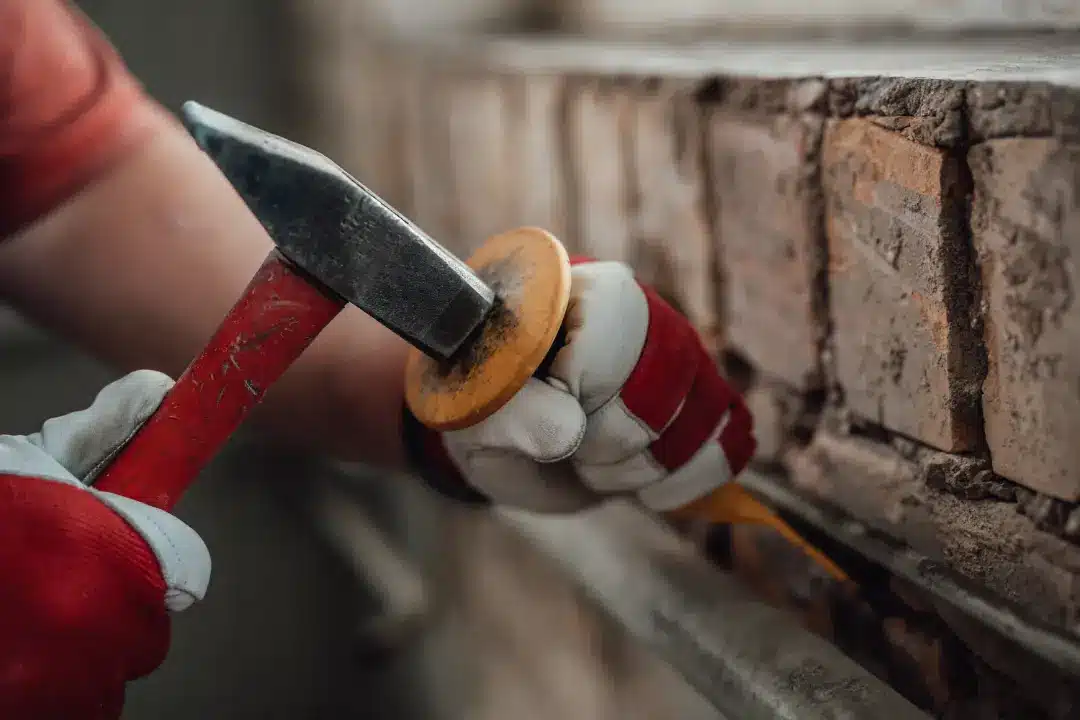 Person using a hammer and chisel to remove old mortar from brickwork, illustrating the process of repointing for property restoration.