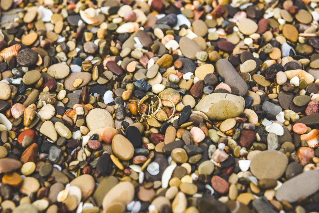 Two gold wedding rings nestled among a variety of smooth pebbles on a beach, highlighting the contrast between the rings and the natural stones.